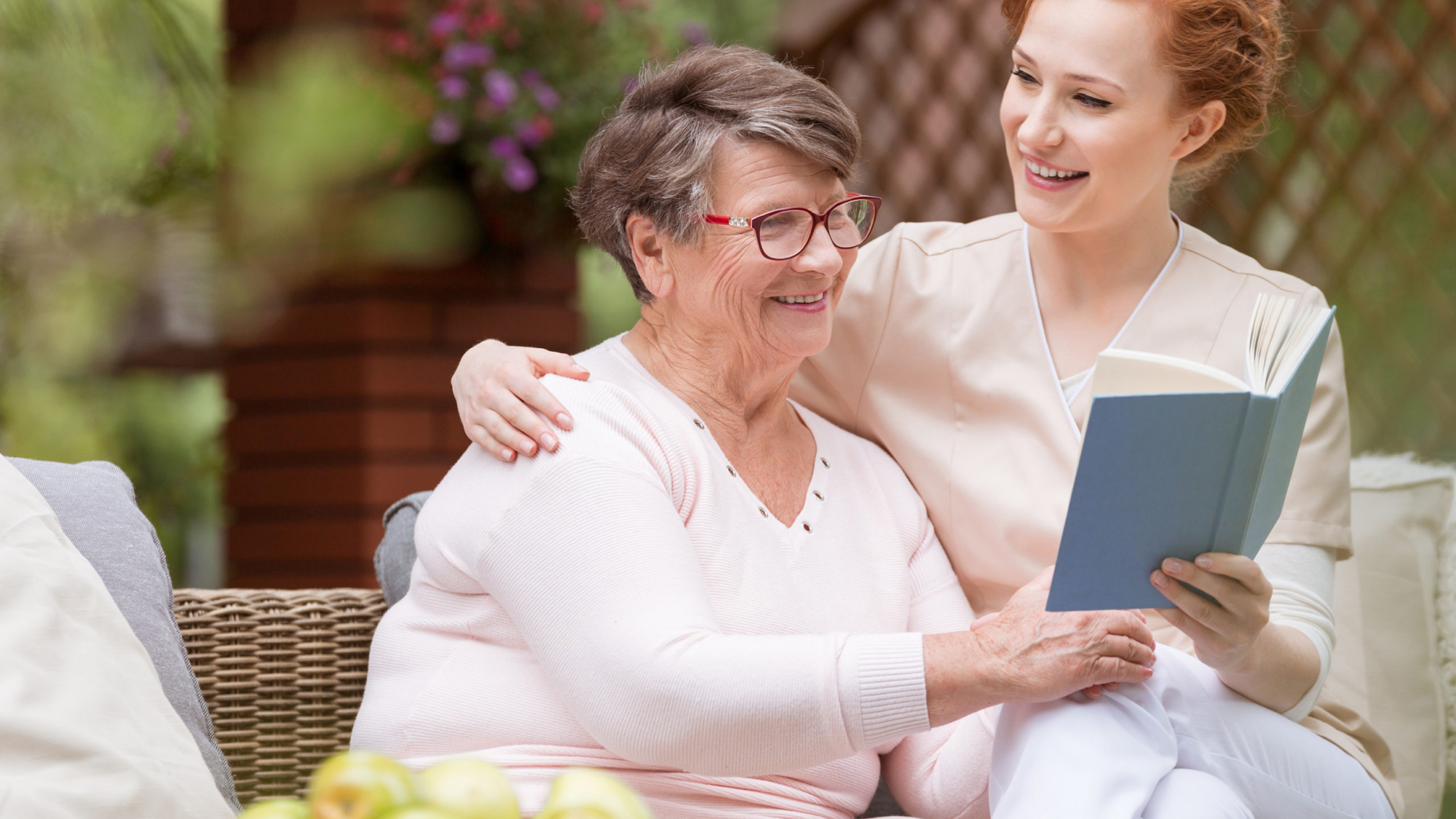 Cheerful senior woman with her tender caretaker reading a book together while relaxing outside. Close relationship.