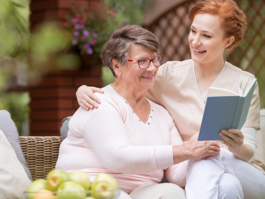 Cheerful senior woman with her tender caretaker reading a book together while relaxing outside. Close relationship.