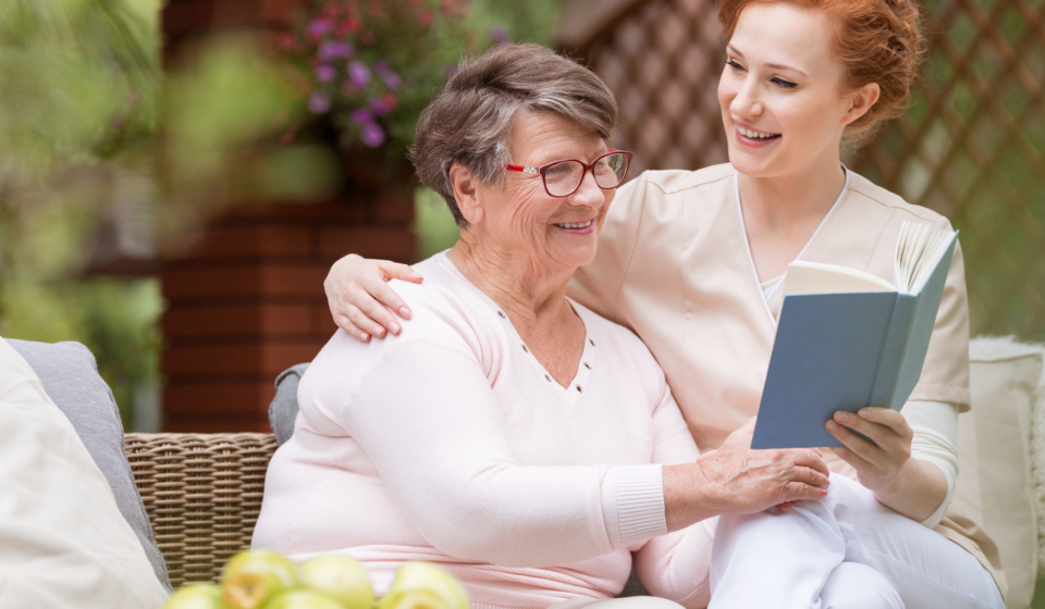 Cheerful senior woman with her tender caretaker reading a book together while relaxing outside. Close relationship.